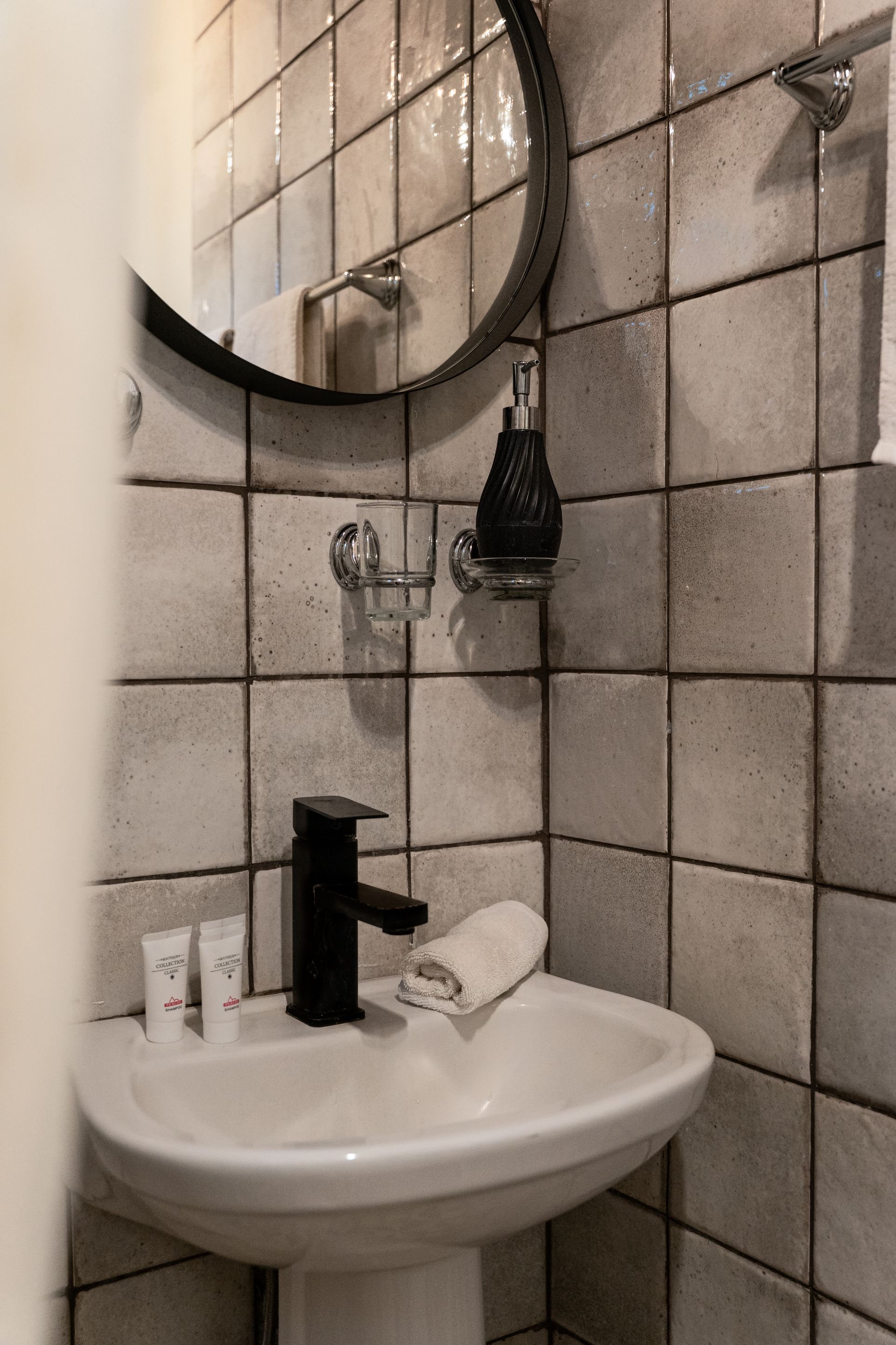 Small white sink with black faucet, round mirror, and tiled bathroom wall with shelves and toiletries.
