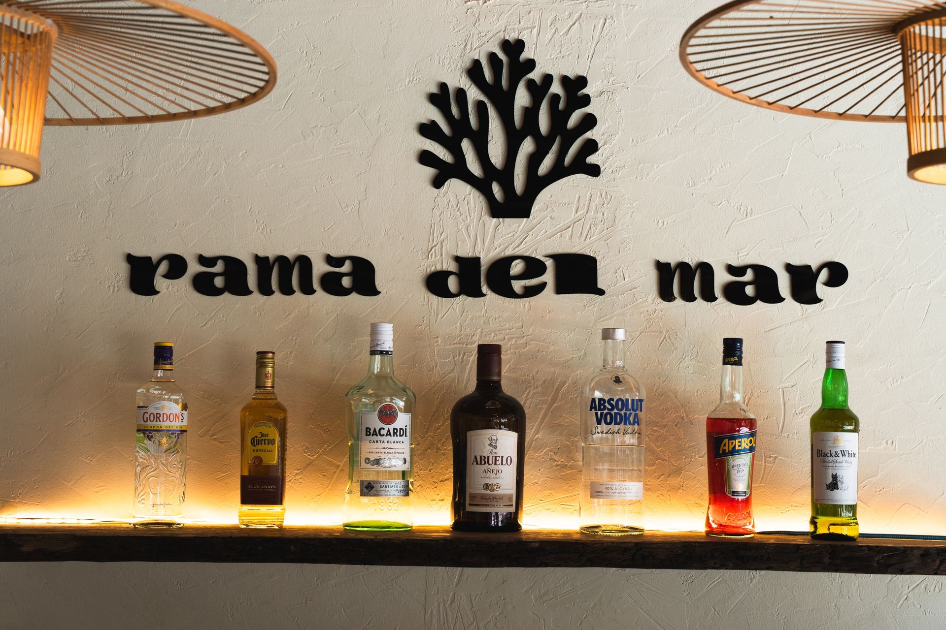 Bar sign reading “rama del mar” with bottles on a lit shelf beneath hanging lamps