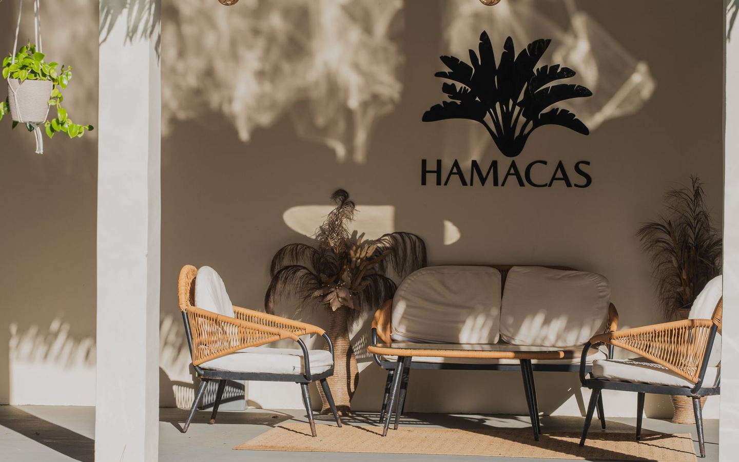 Outdoor patio with wicker chairs, cushioned sofa, and a wall sign reading “HAMACAS” under a tree shadow