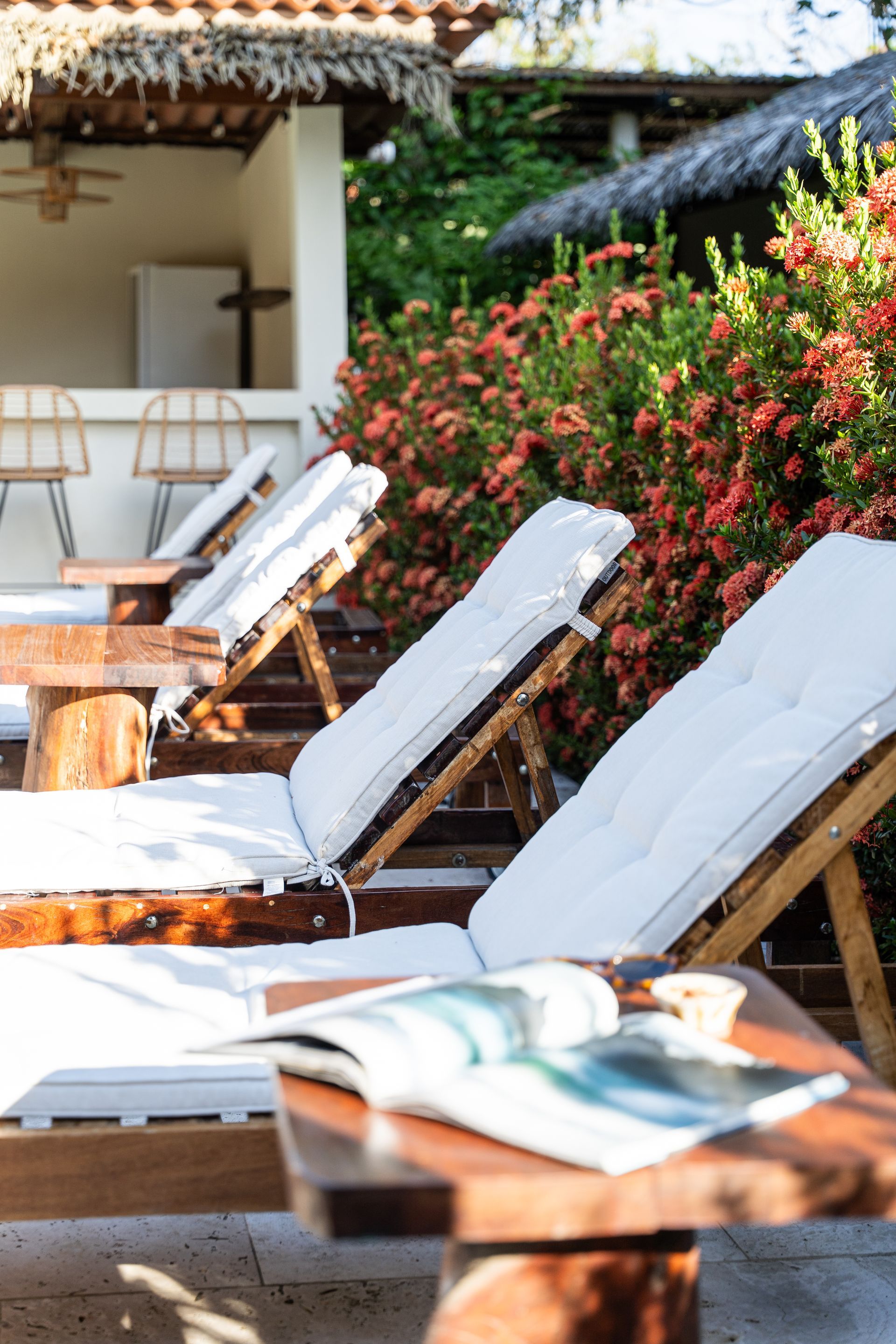 White lounge chairs beside a flower-lined patio by the pool, with sunlit resort seating in the background
