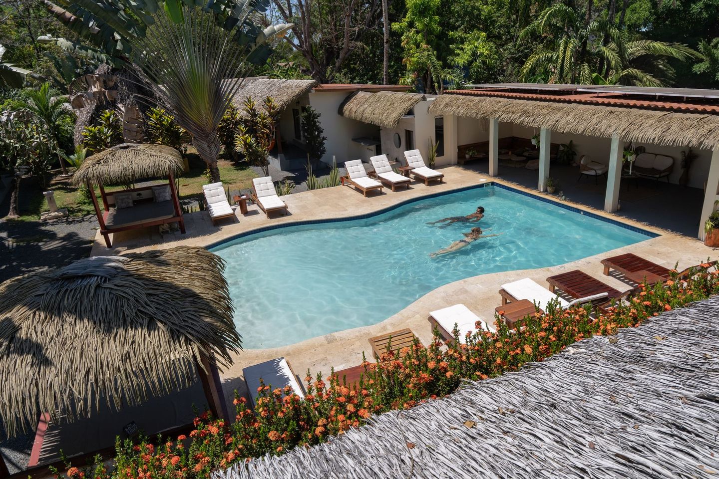 Resort-style pool with lounge chairs, thatched cabanas, and a person swimming in clear blue water