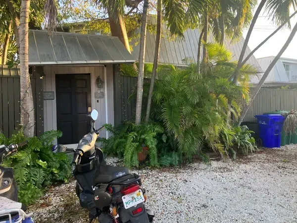 Scooter parked outside a small house with tropical plants, palm trees, and blue trash bins along a gravel driveway.