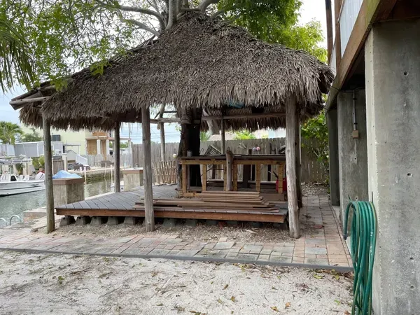 Thatched-roof patio with wooden deck and tables beside a concrete building, with a coiled green hose nearby.