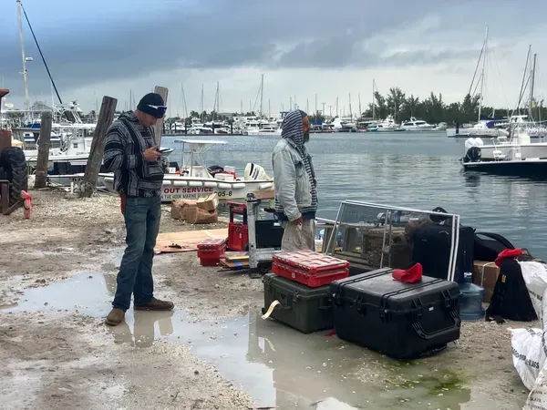Two people in winter gear on a snowy marina dock beside stacked gear and boats on calm water