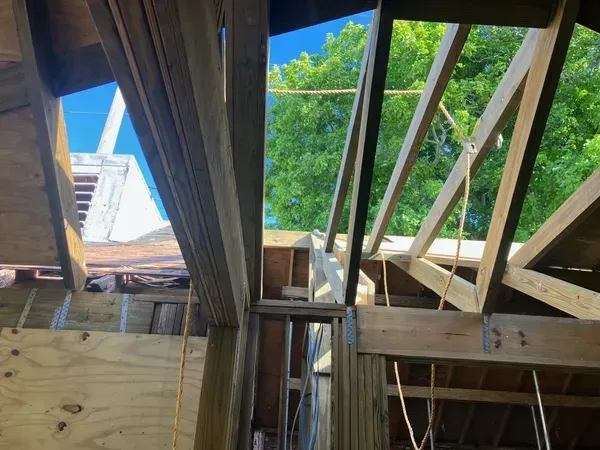View through exposed roof framing to a blue sky, leafy tree, and neighboring building.