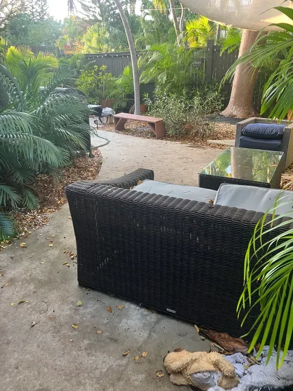 Tropical patio with black wicker seating, sandy path, lush greenery, and a small tan dog resting in front.