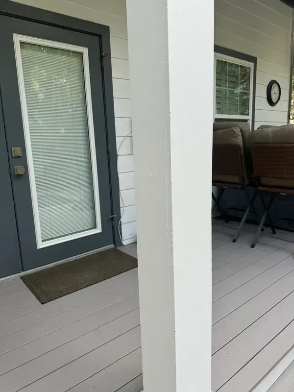 Front porch with dark gray door, white column, and outdoor seating beside a window.