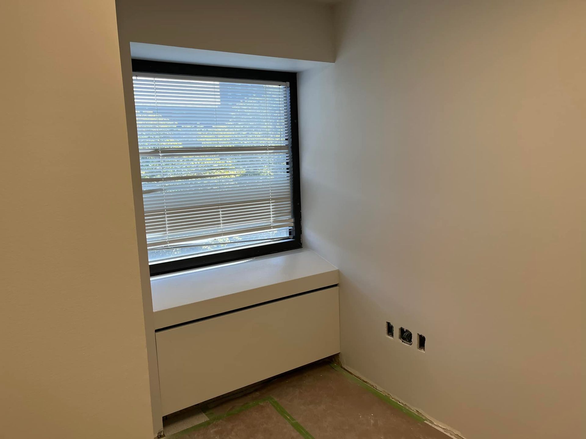 Empty white room with a window, radiator cover, and unfinished floor with green tape along the baseboards