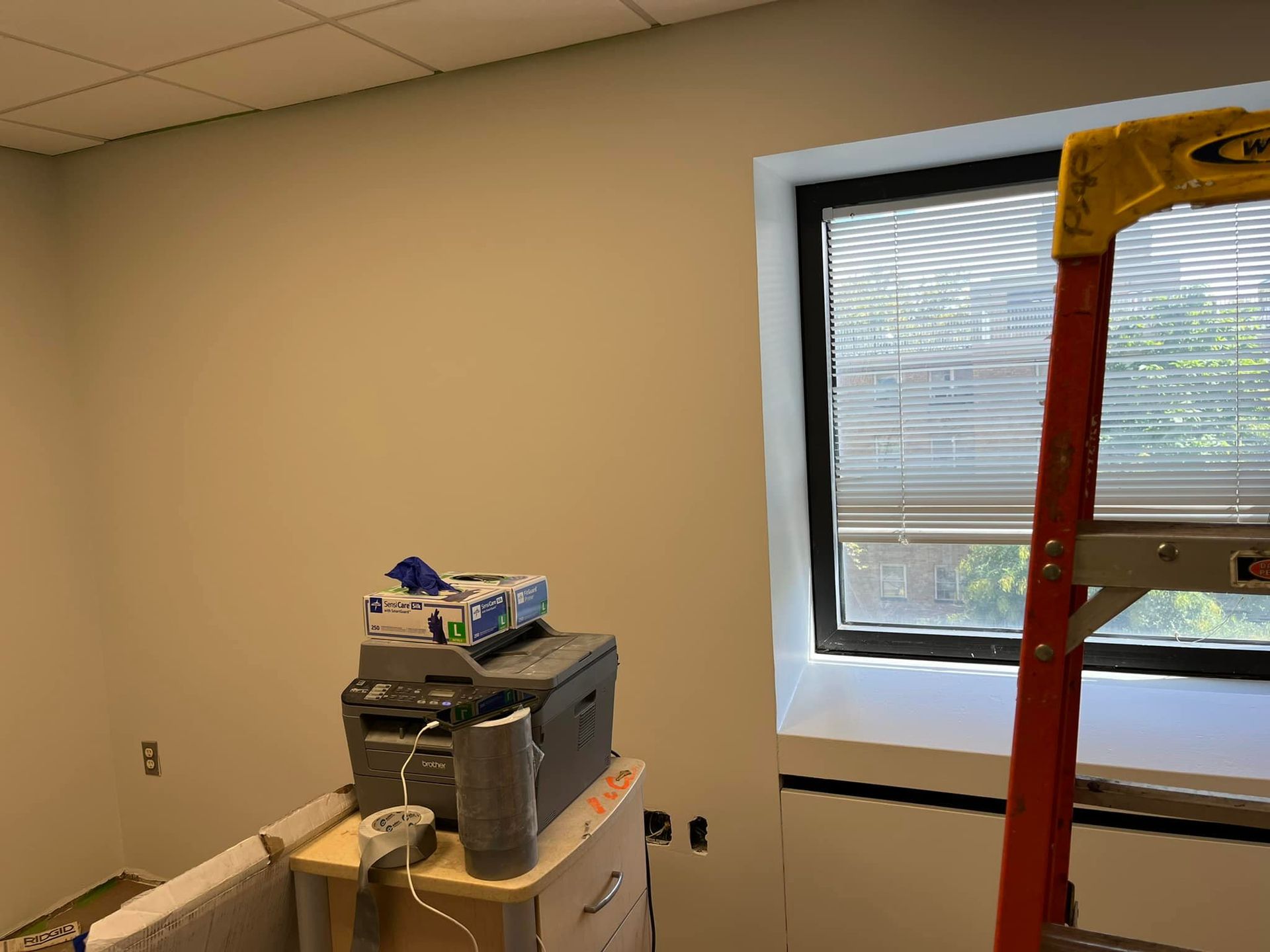 Room with beige walls, window blinds, stacked equipment on a cart, and a red ladder near the window.