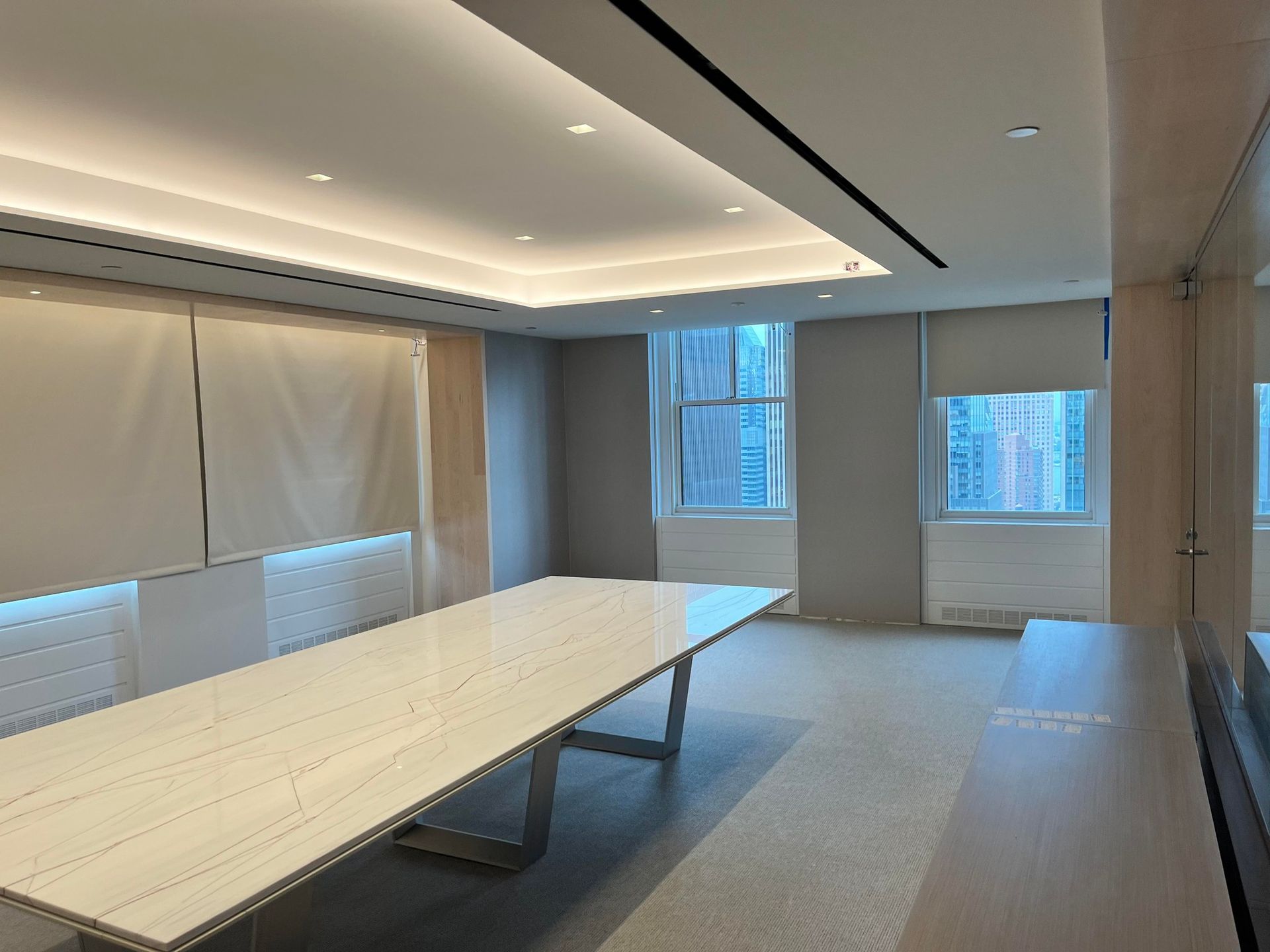 Empty conference room with long white table, recessed ceiling lights, and large windows with blinds.