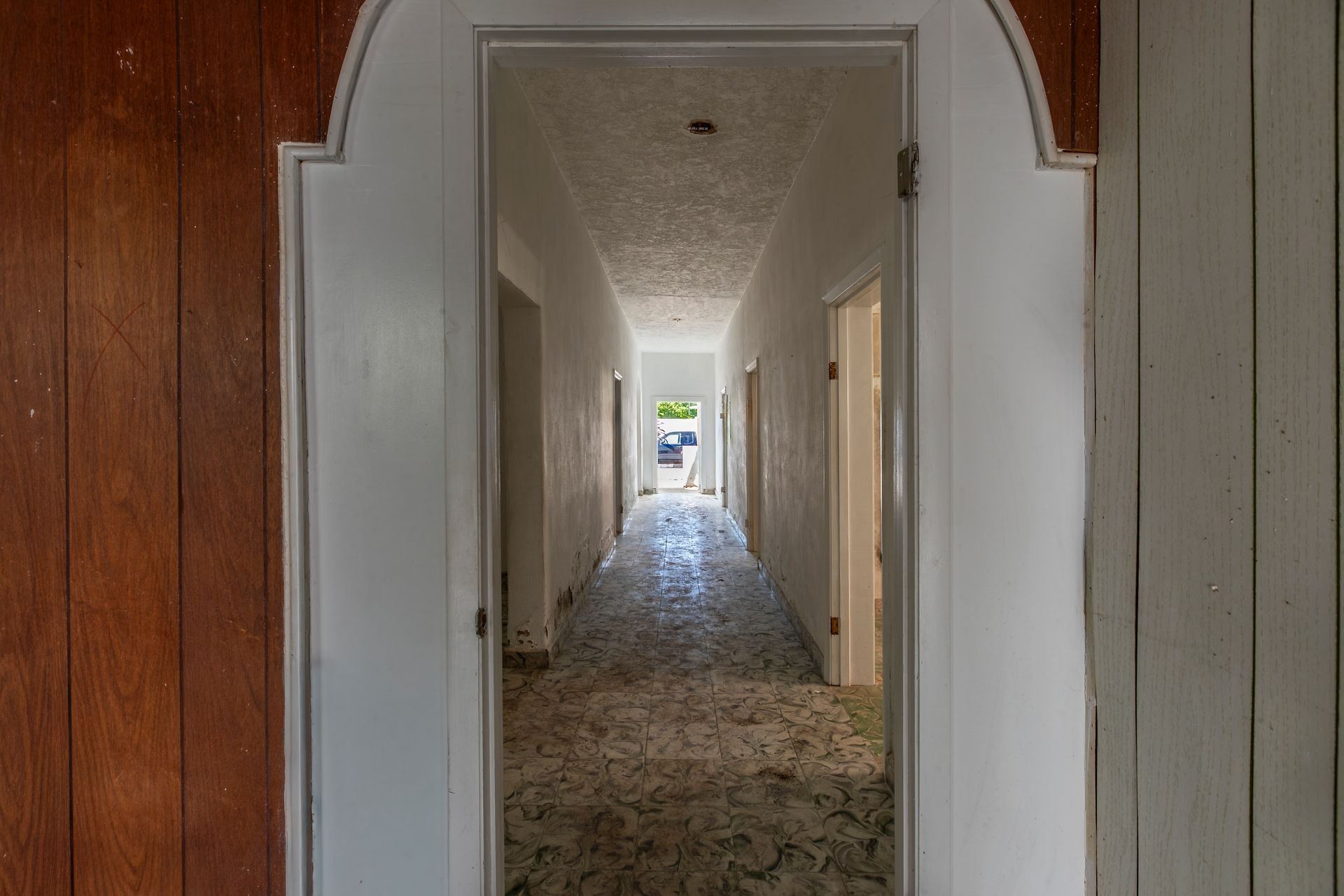 Narrow hallway with peeling walls and patterned carpet, leading to a bright doorway at the far end