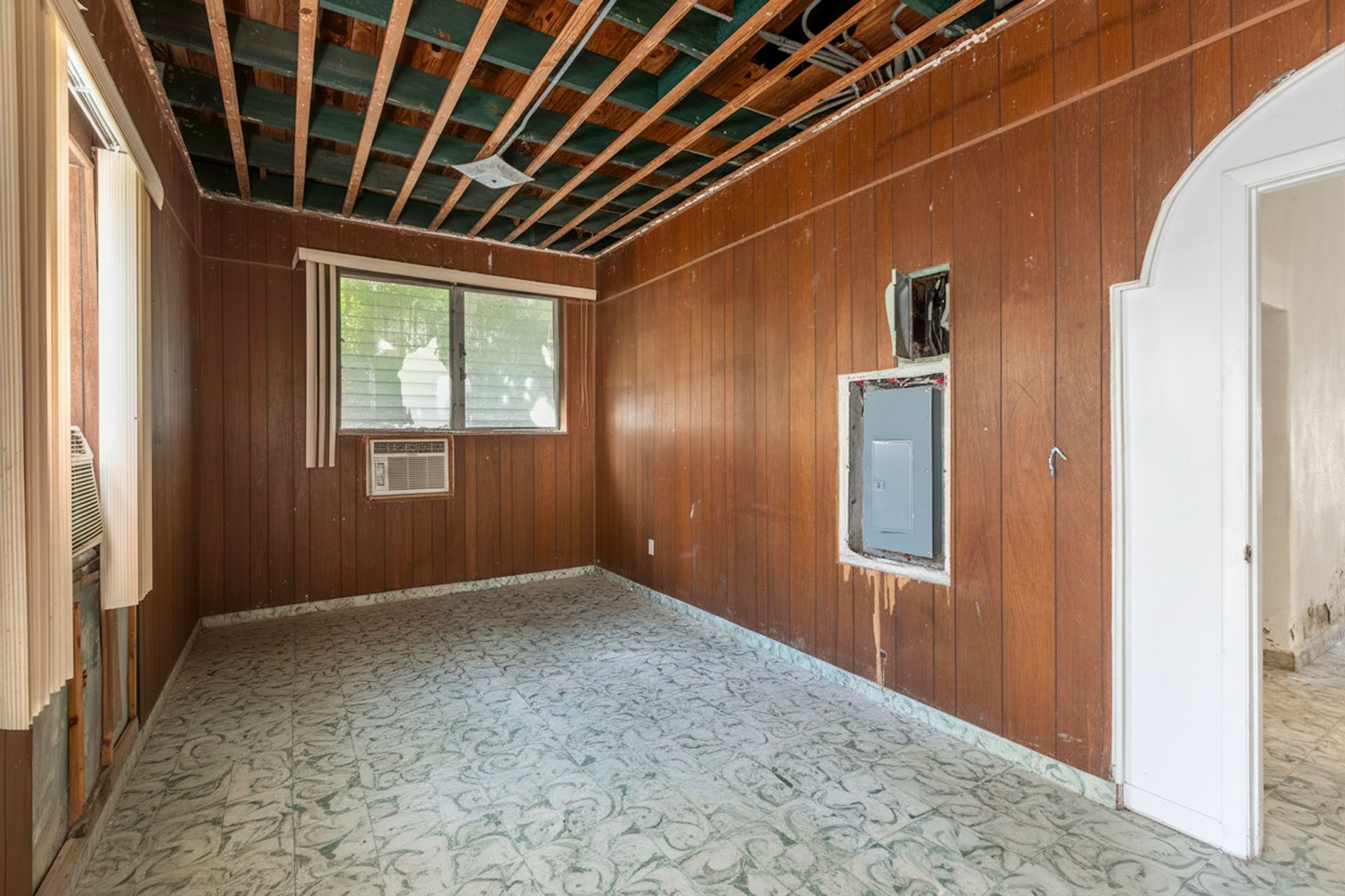 Empty wood-paneled room with exposed ceiling beams, patterned floor, window, and arched doorway