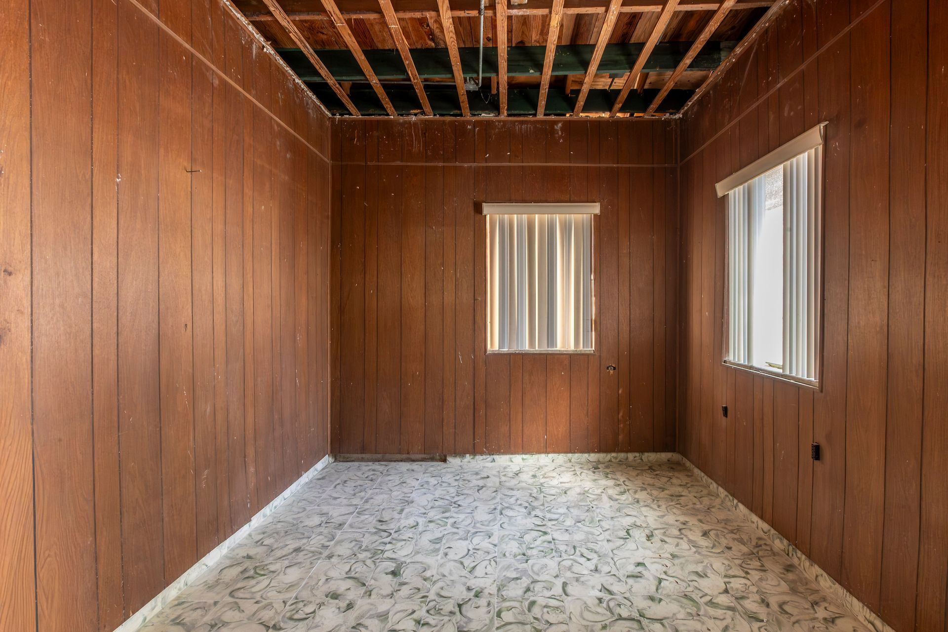 Empty unfinished room with wood-paneled walls, exposed ceiling joists, and two windows.