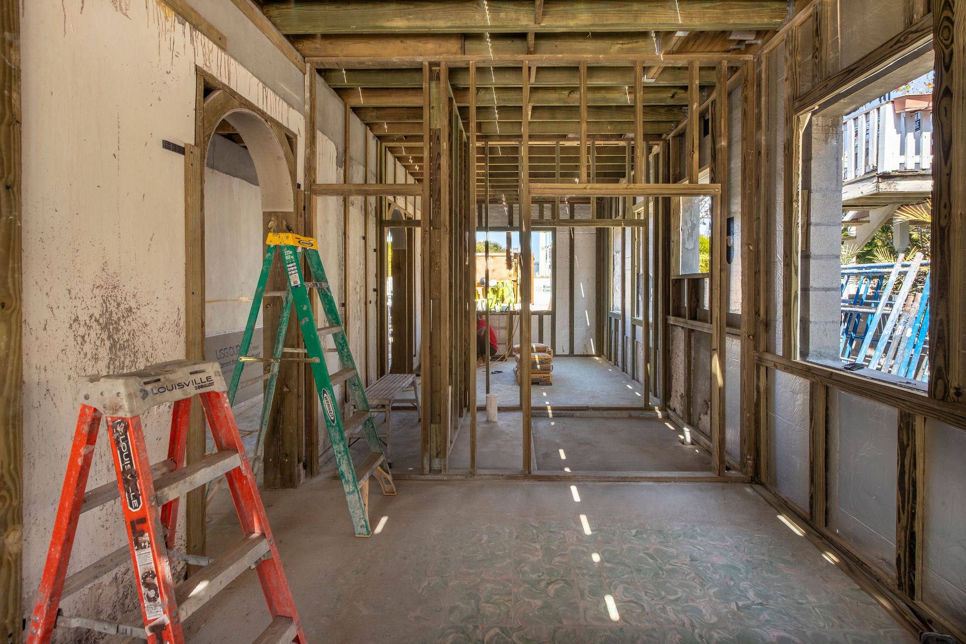 Partially framed room under renovation with ladders, exposed studs, and sunlight through window openings