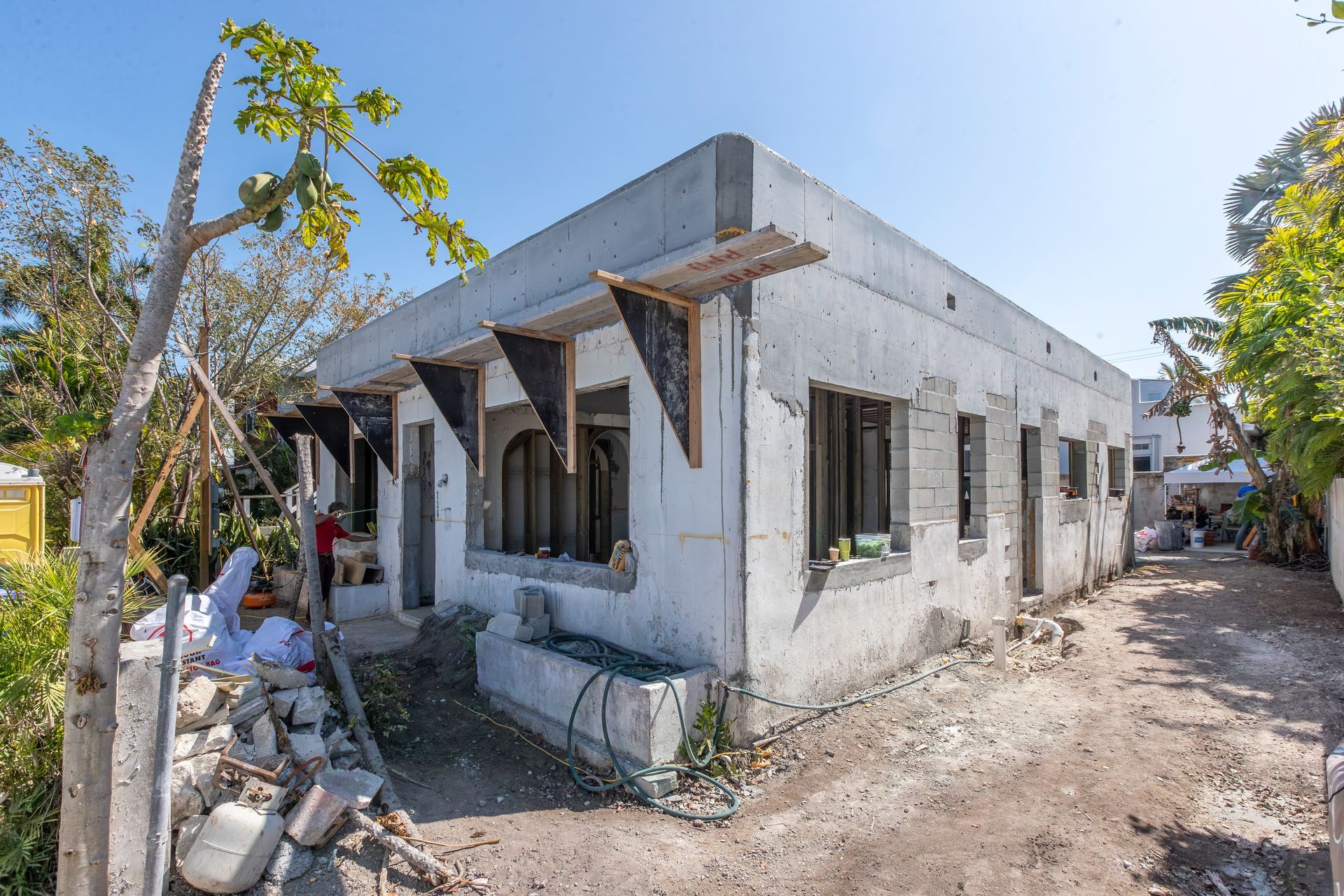 Unfinished concrete house with windows and wooden supports along a dirt path under a clear blue sky