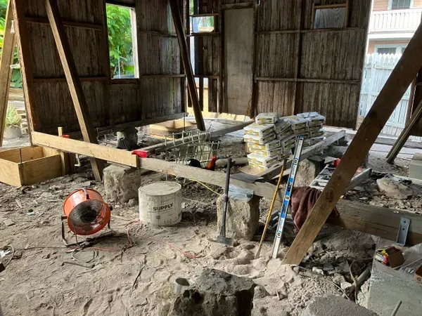 Partially built wooden shack with stacks of cinder blocks, tools, and a cement mixer on a dirt floor.