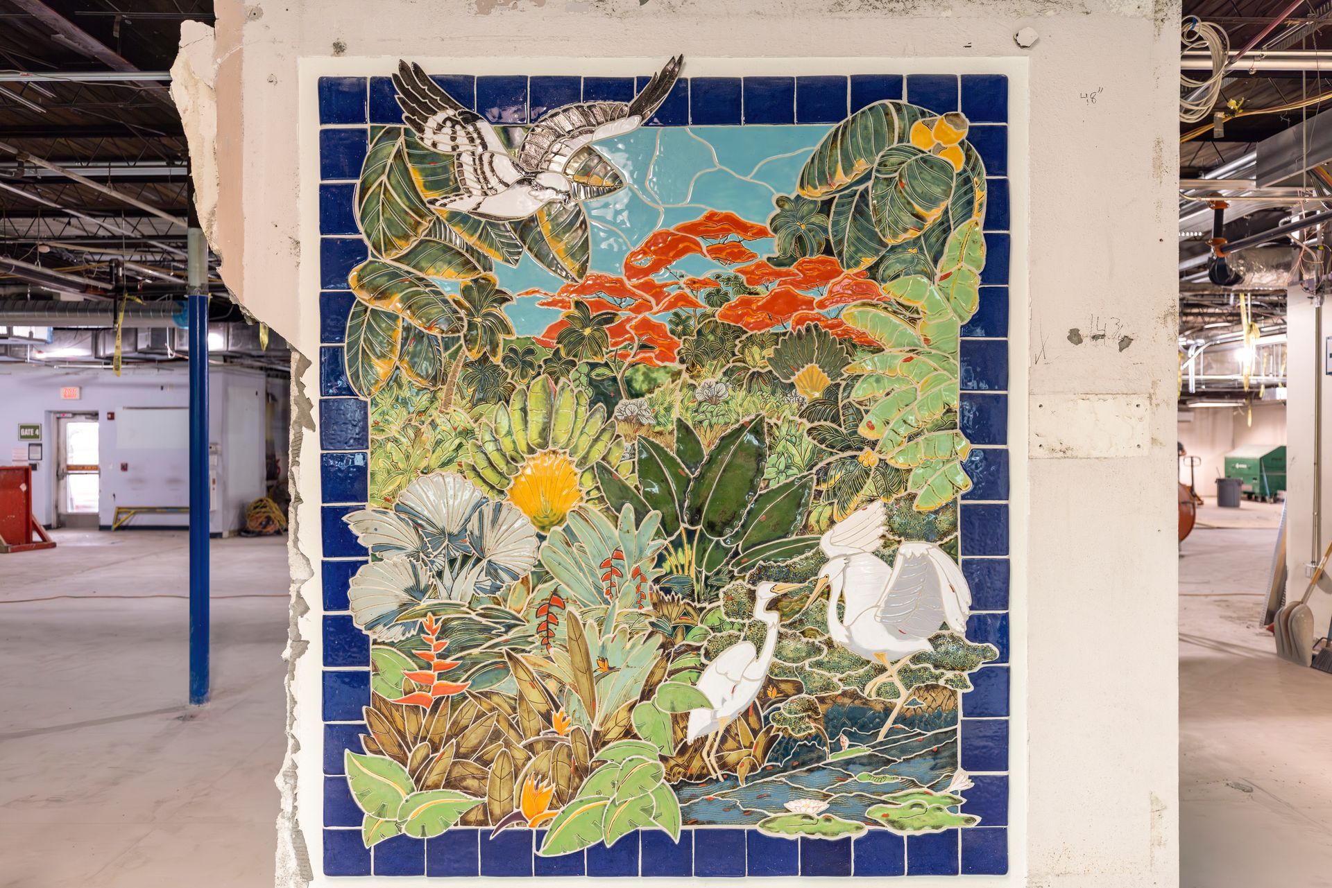 Colorful mosaic mural of birds, plants, and fruit on a concrete pillar in an industrial space