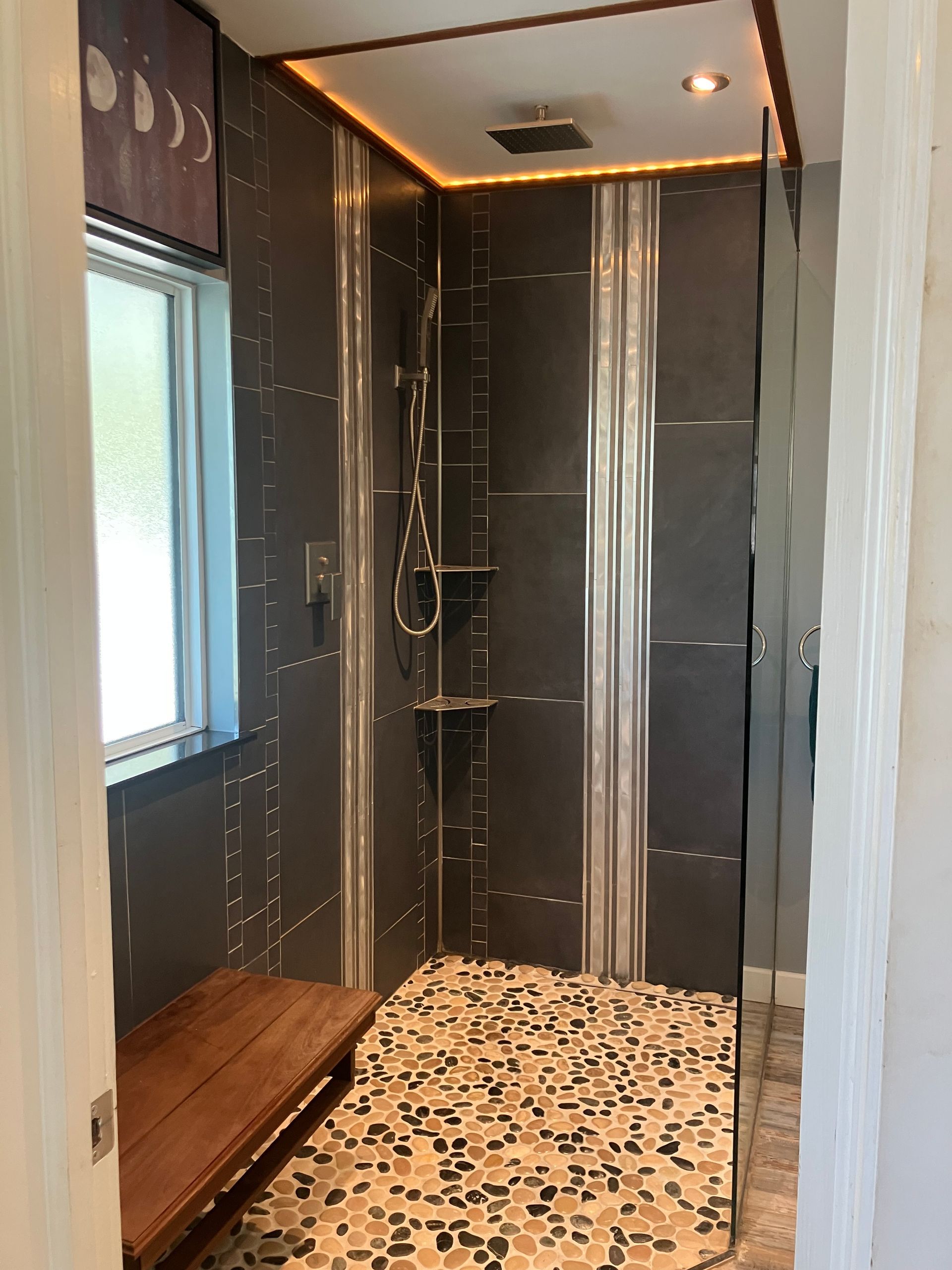 Narrow modern shower with dark tile walls, patterned floor, wooden bench, and window