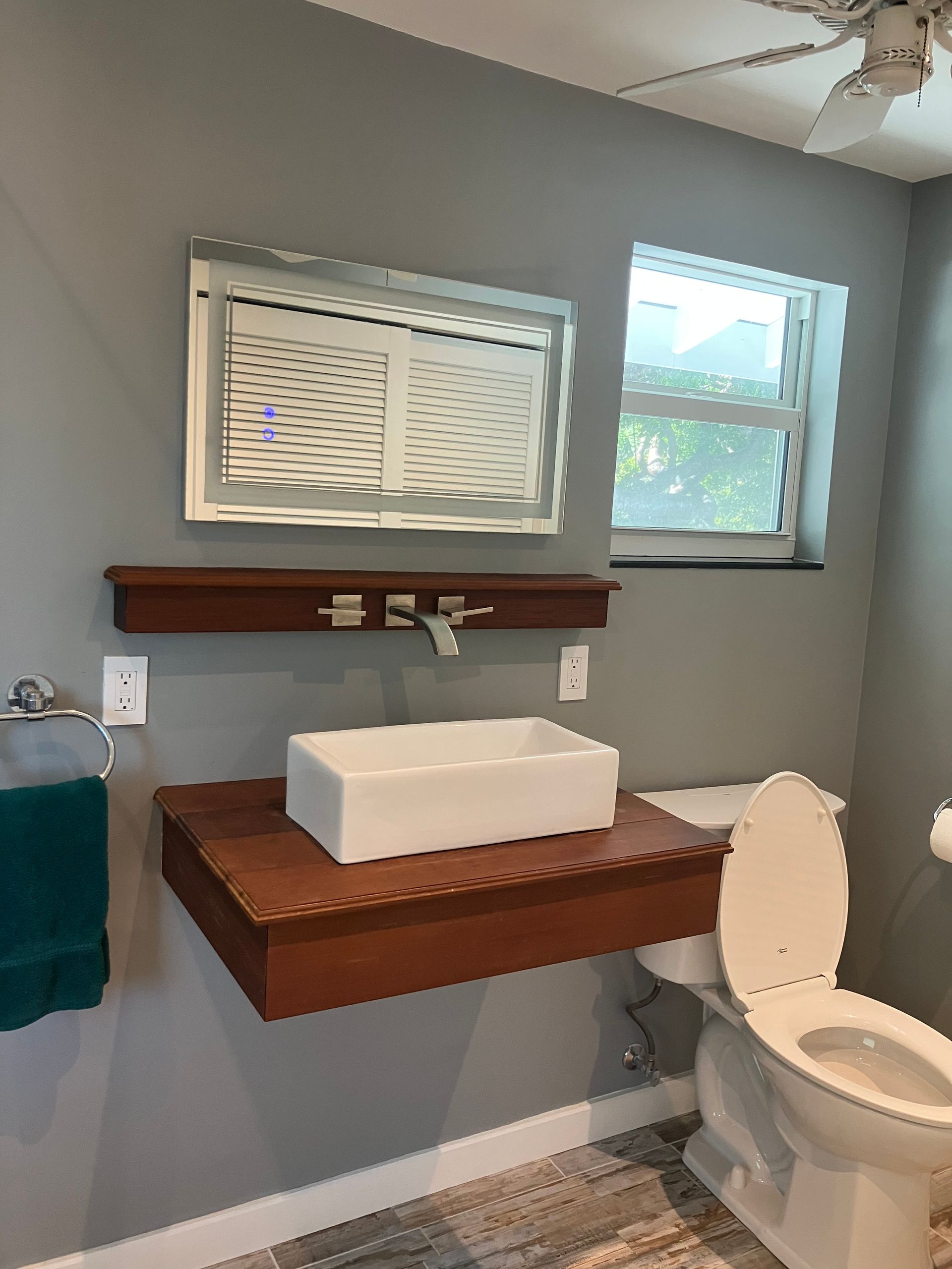 Bathroom with floating wood vanity, white vessel sink, mirror, window, towel, and toilet.