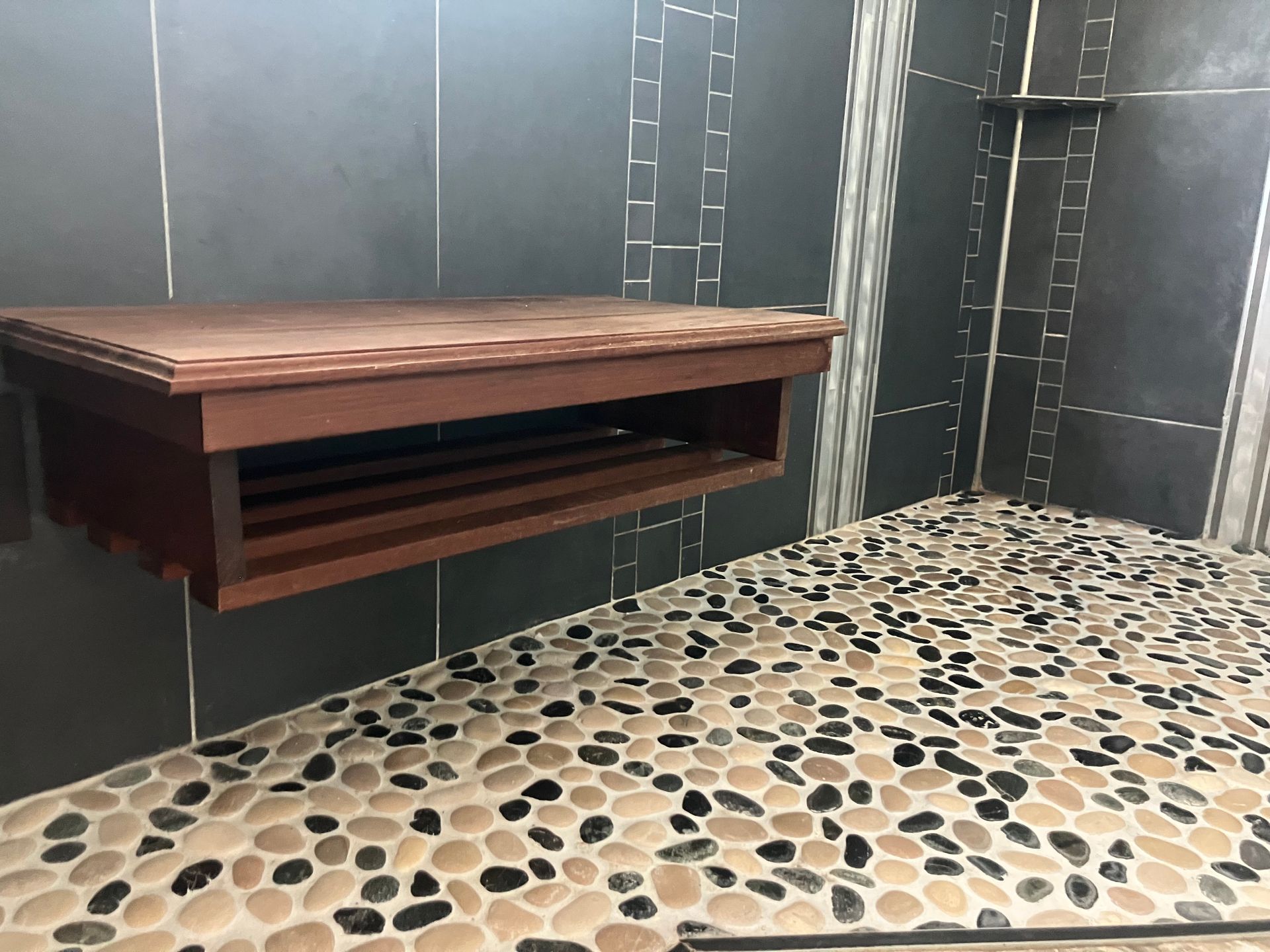 Modern tiled shower with pebble floor and wooden wall shelf