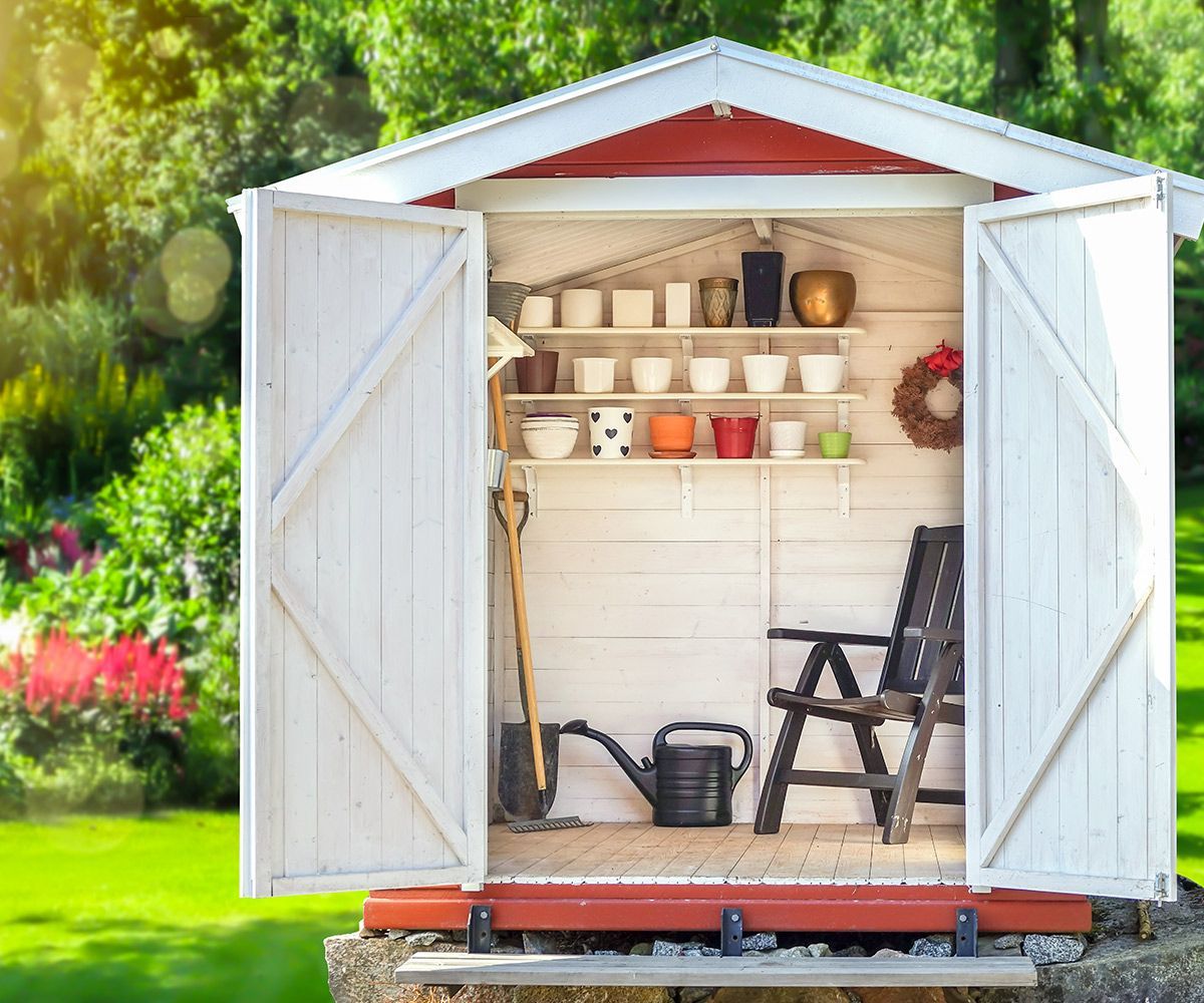 Open garden shed with white doors, shelves of pots, ladder, watering can, and yard tools inside.