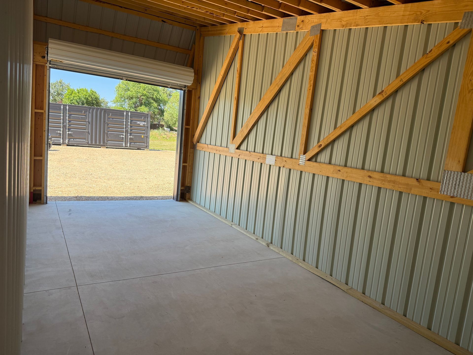 Empty covered carport with concrete floor, corrugated walls, and open view to a sunny yard.