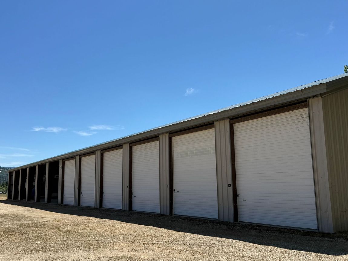 Row of white storage unit doors beneath a slanted roof on a gravel lot under a clear blue sky