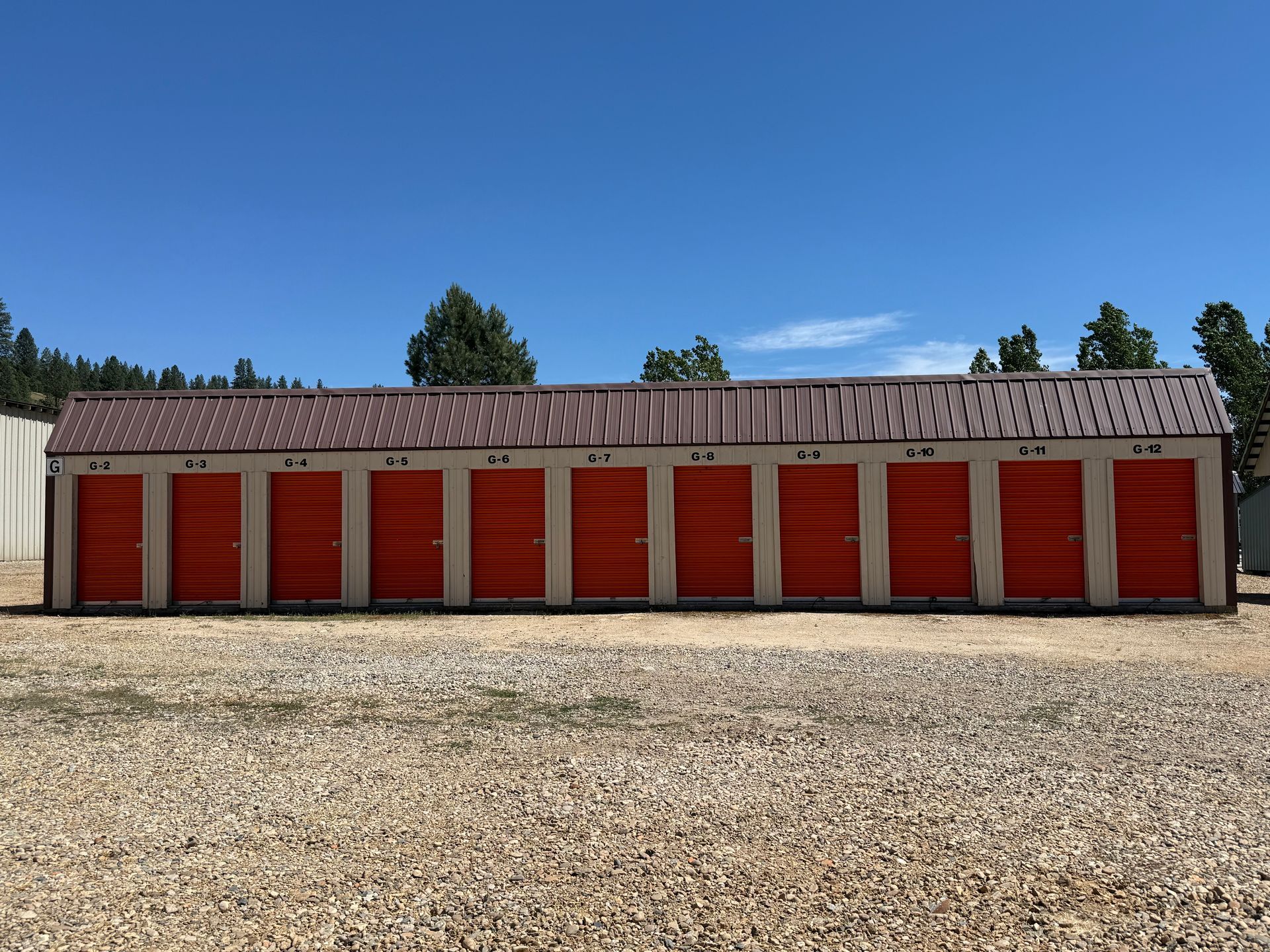 Row of red storage unit doors with a gray roof on a gravel lot under a clear blue sky