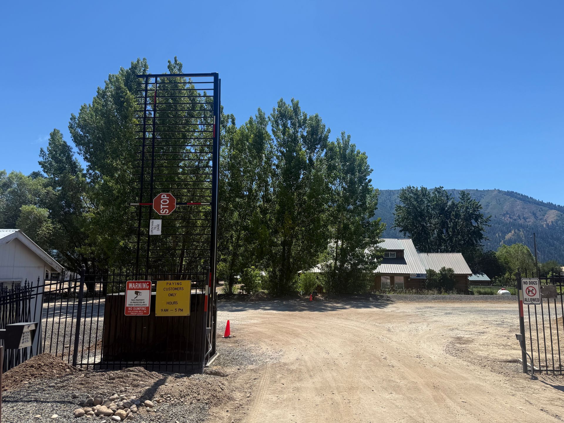 Open gate to a dirt lot with trees, white buildings, and a mountain under a clear blue sky.