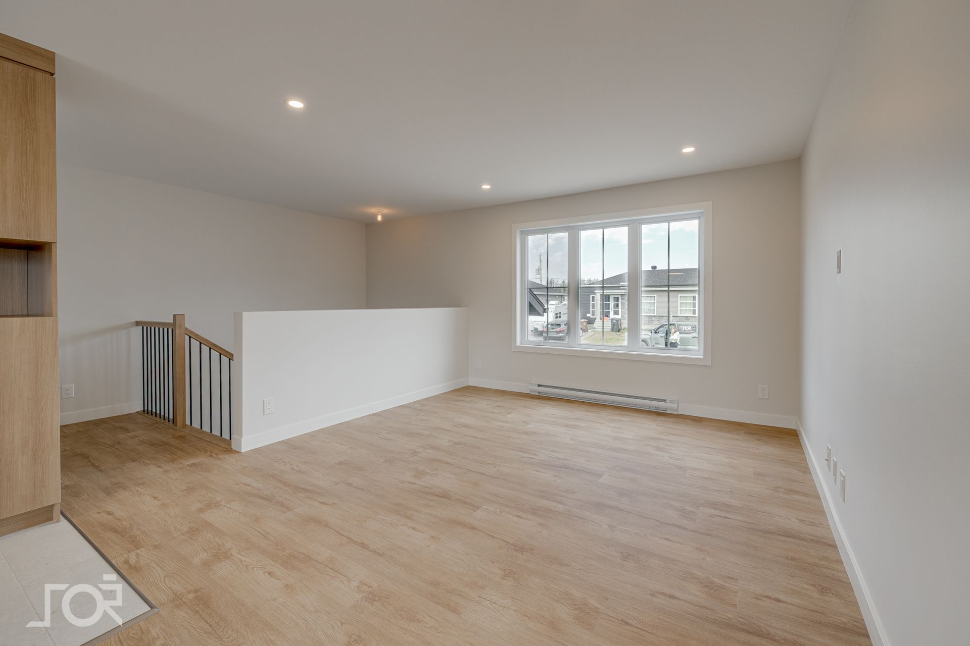 Pièce vide avec parquet, grande fenêtre et escalier. Placards en bois clair sur la gauche.