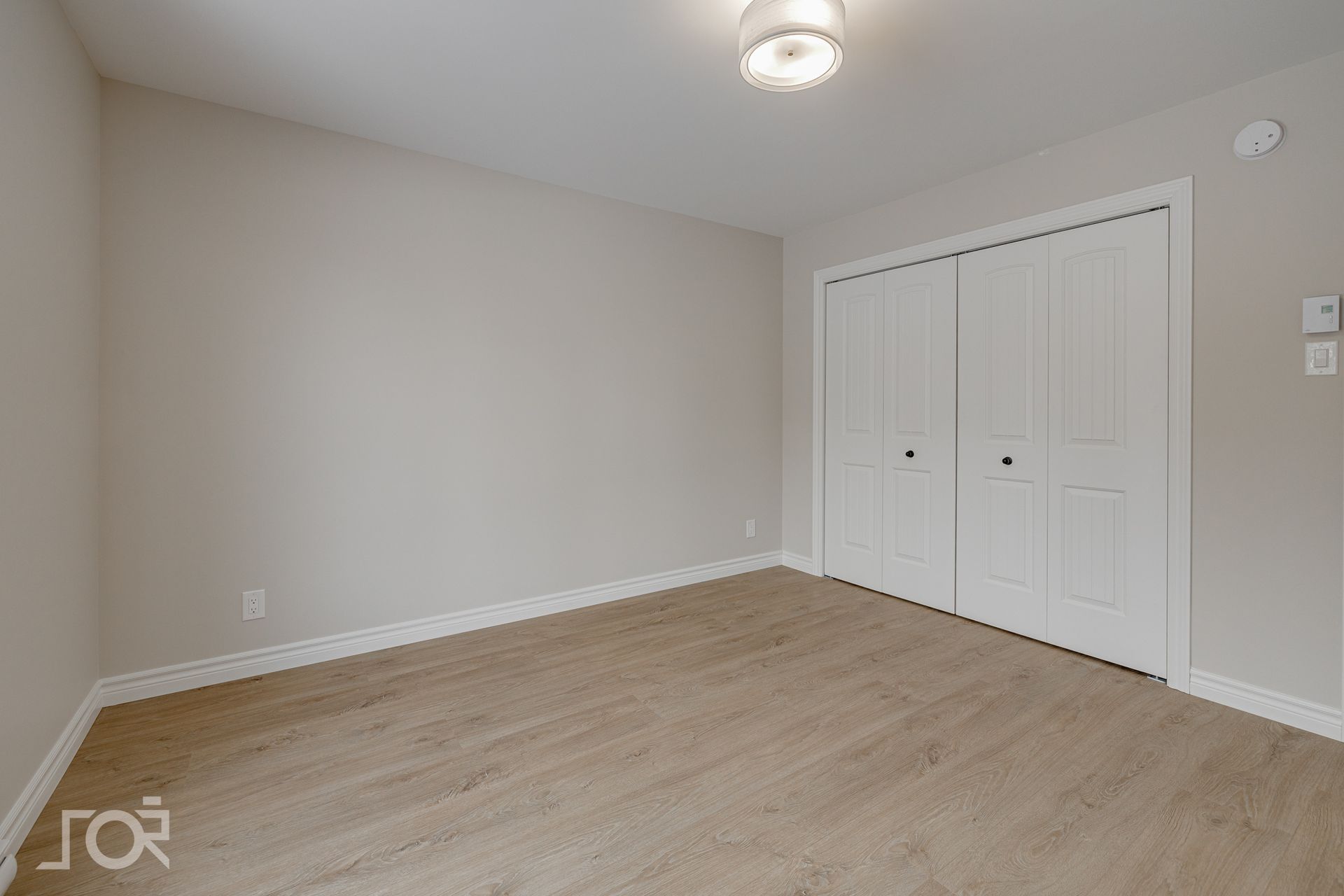 Chambre vide avec murs beige clair, placard blanc à deux portes et sol imitation bois.