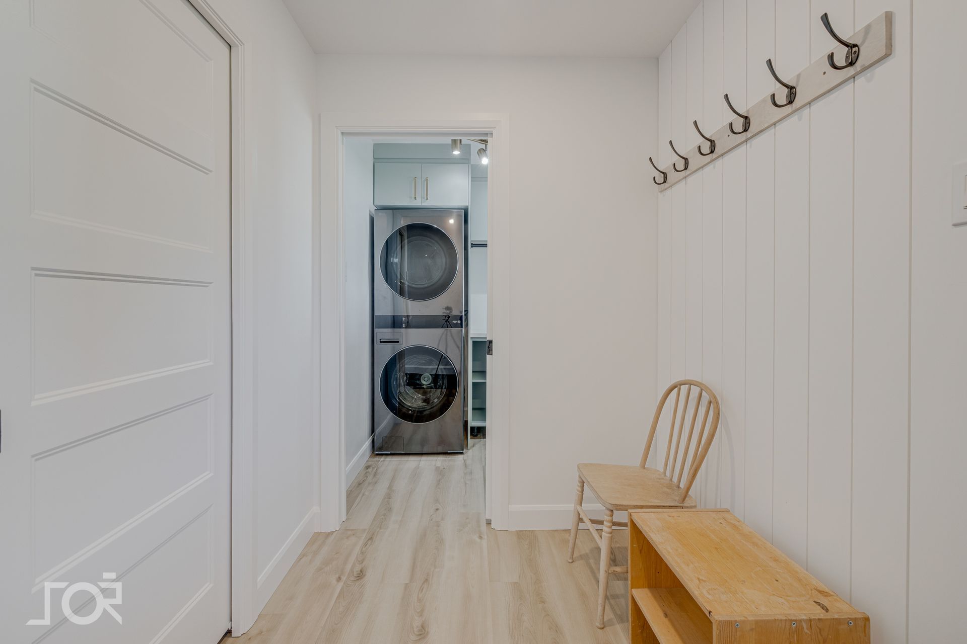 Couloir blanc avec machines à laver superposées, banc en bois, chaise et porte-manteau.