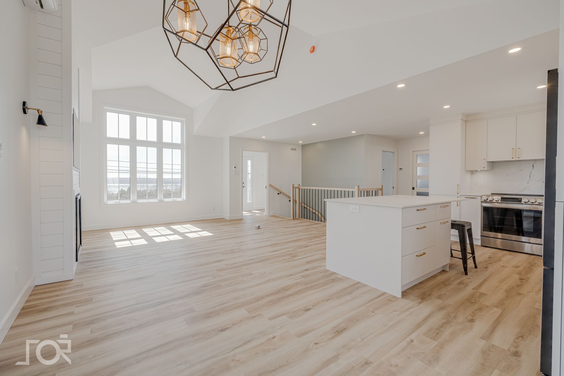 Espace de vie lumineux et ouvert avec îlot de cuisine, grande fenêtre, parquet et luminaire géométrique.