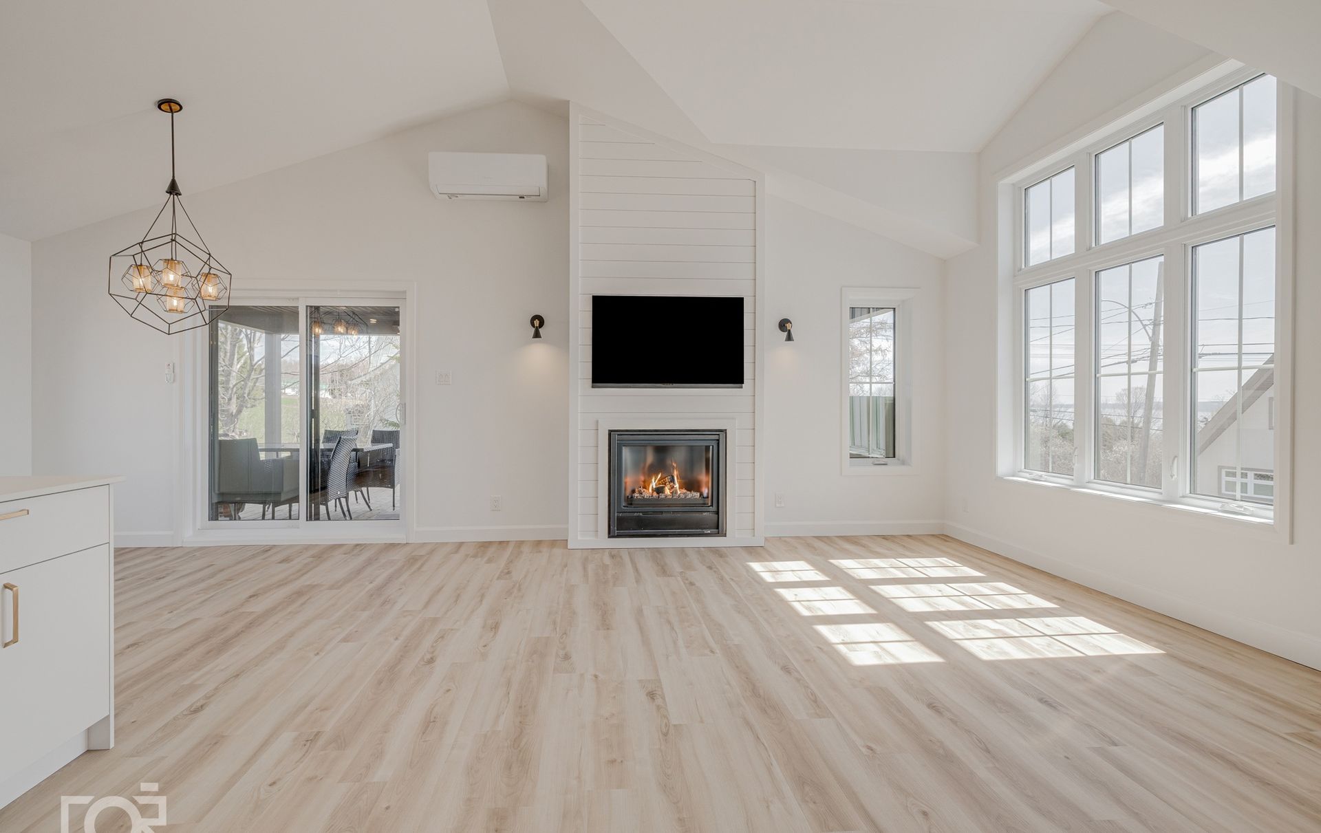 Salon lumineux et vide avec cheminée, télévision et grandes fenêtres. Parquet clair, murs blancs.