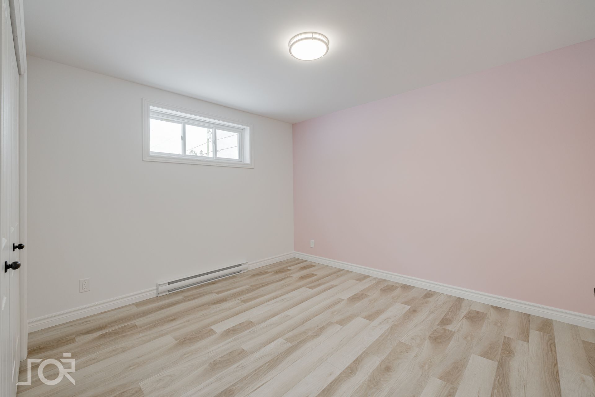 Pièce vide avec parquet clair, murs blancs et roses et une petite fenêtre.