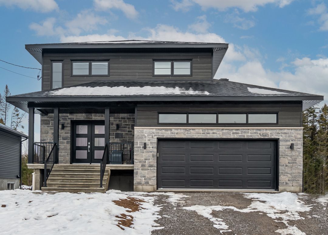 Une grande maison avec une porte de garage noire est assise au sommet d'une colline couverte de neige.