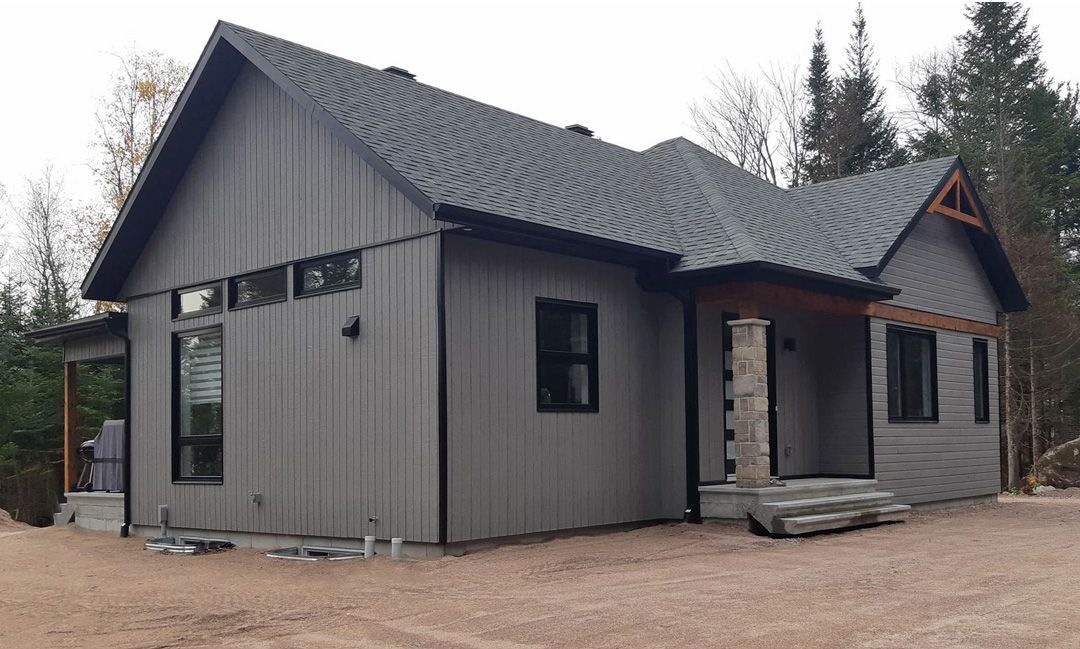 Une maison grise avec un toit noir est assise au sommet d'un champ de terre.