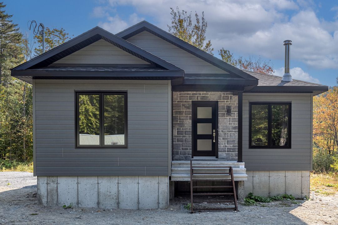 Une petite maison grise avec une porte et des fenêtres noires est assise sur un terrain en gravier.