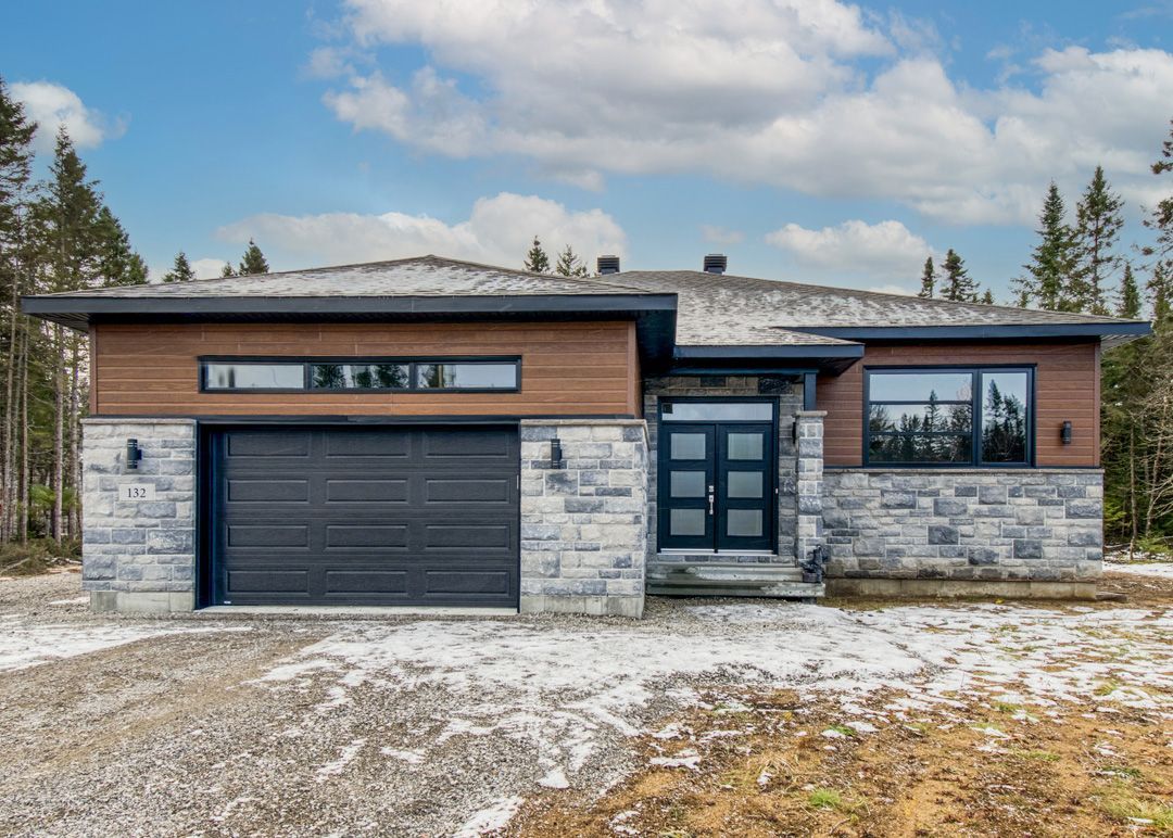 Une maison moderne avec une porte de garage noire est assise au sommet d'une colline de terre.