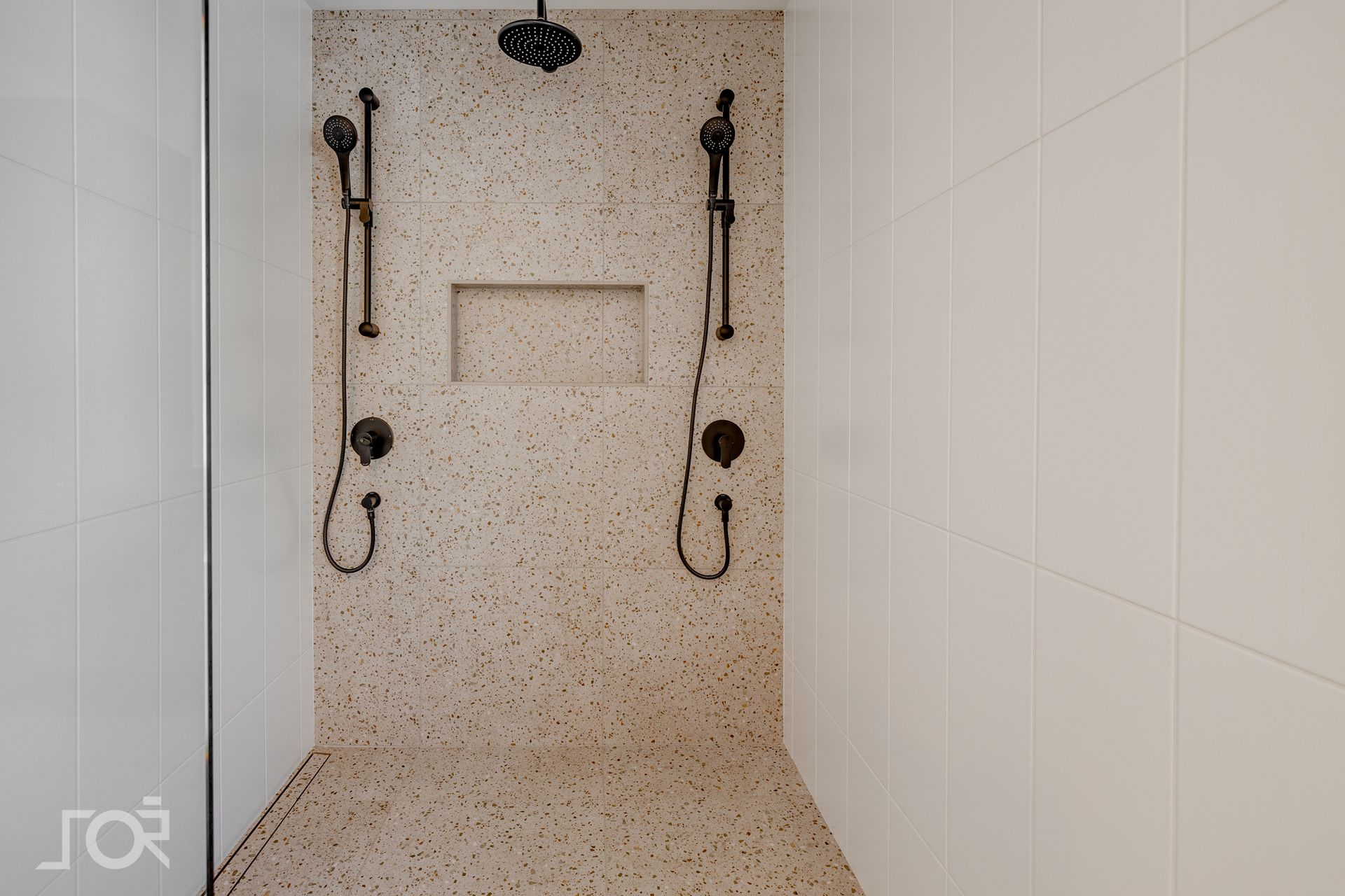 Douche avec murs et sol mouchetés, robinetterie en bronze foncé et murs carrelés blancs.