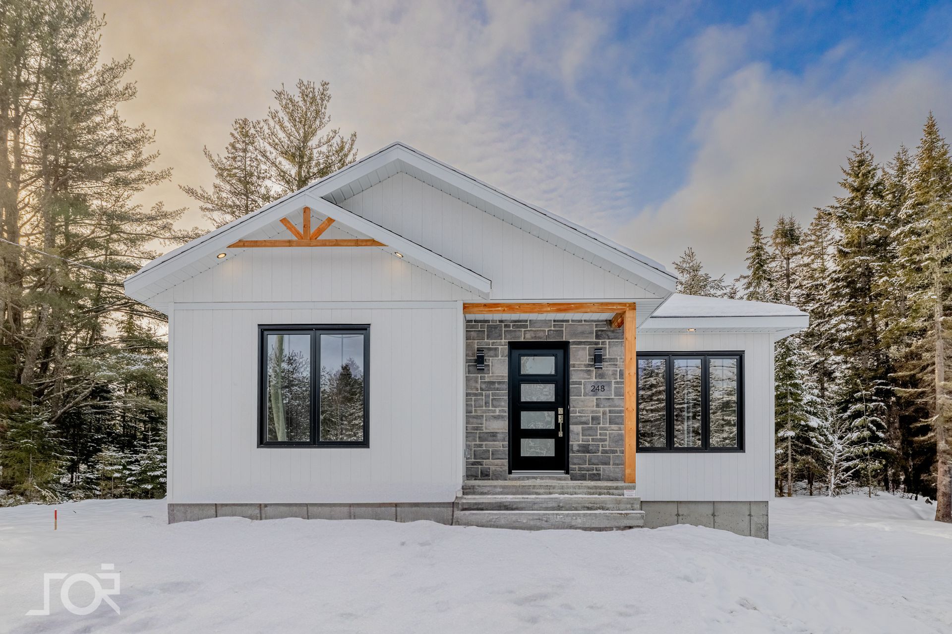 Maison blanche aux fenêtres noires et aux accents de bois, située dans un paysage enneigé.
