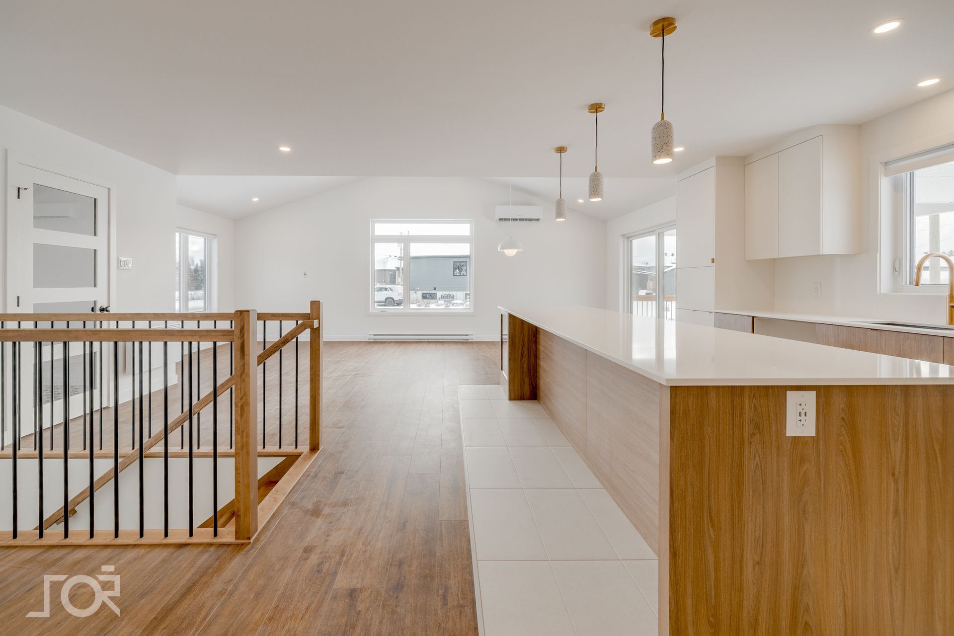 Espace de vie lumineux à aire ouverte avec îlot de cuisine, planchers en bois clair, murs blancs et escalier avec rampes en bois et noires.