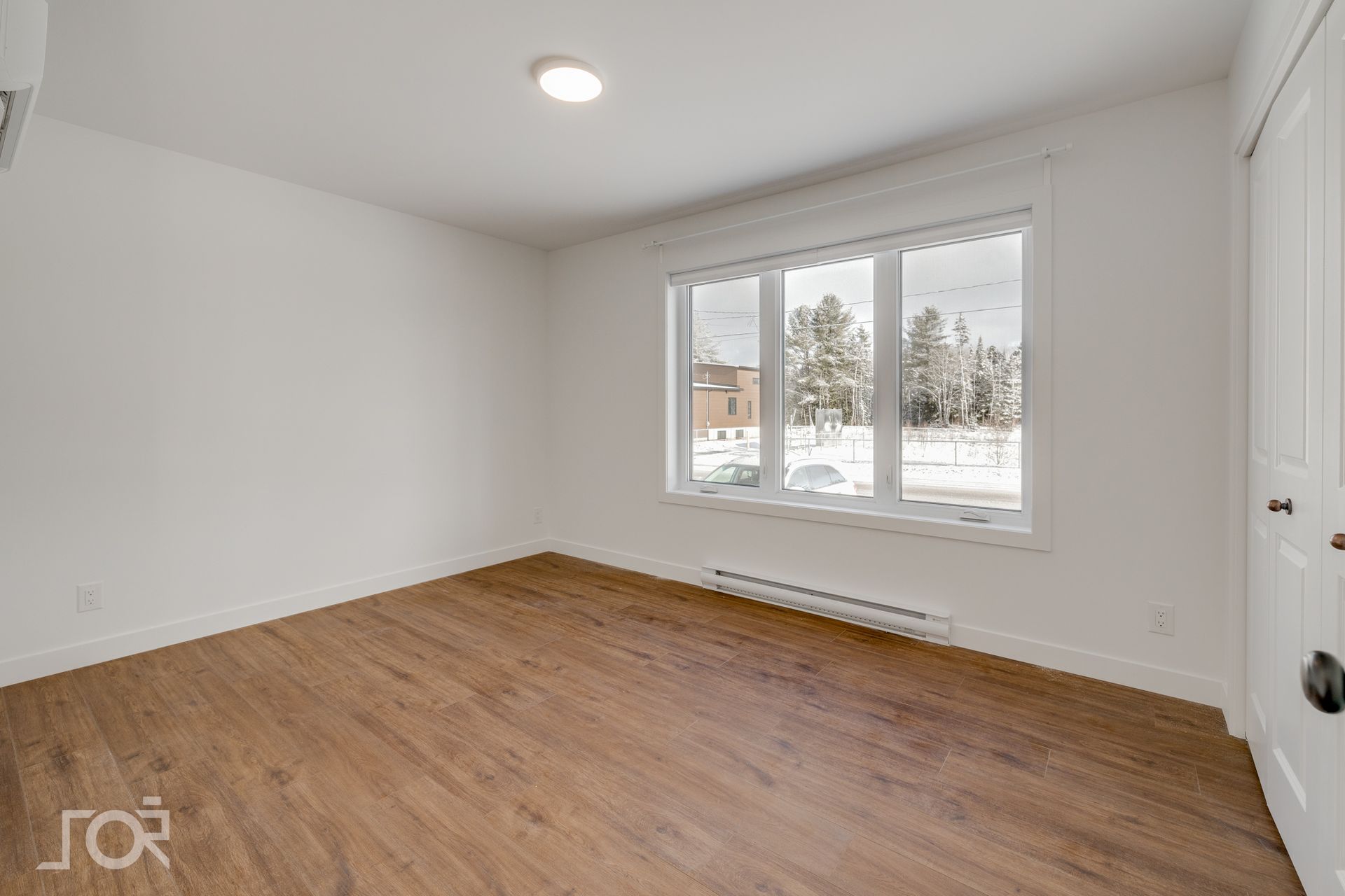 Pièce vide avec parquet, murs blancs et une grande fenêtre donnant sur un paysage extérieur enneigé.