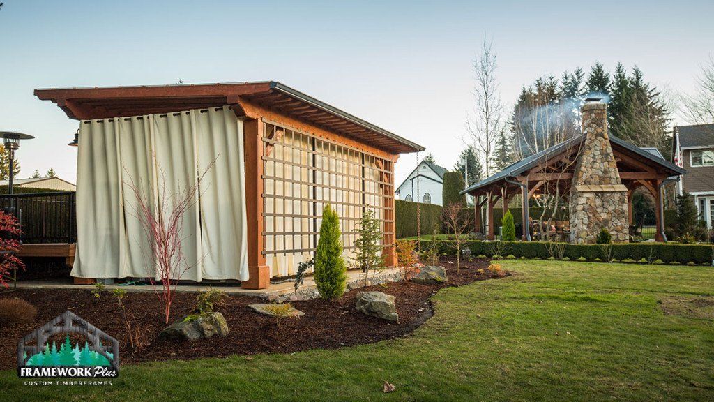 A wooden gazebo with curtains and a stone fireplace in a backyard.