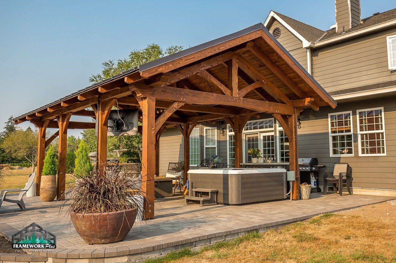 A wooden pavilion with a hot tub underneath it in front of a house.