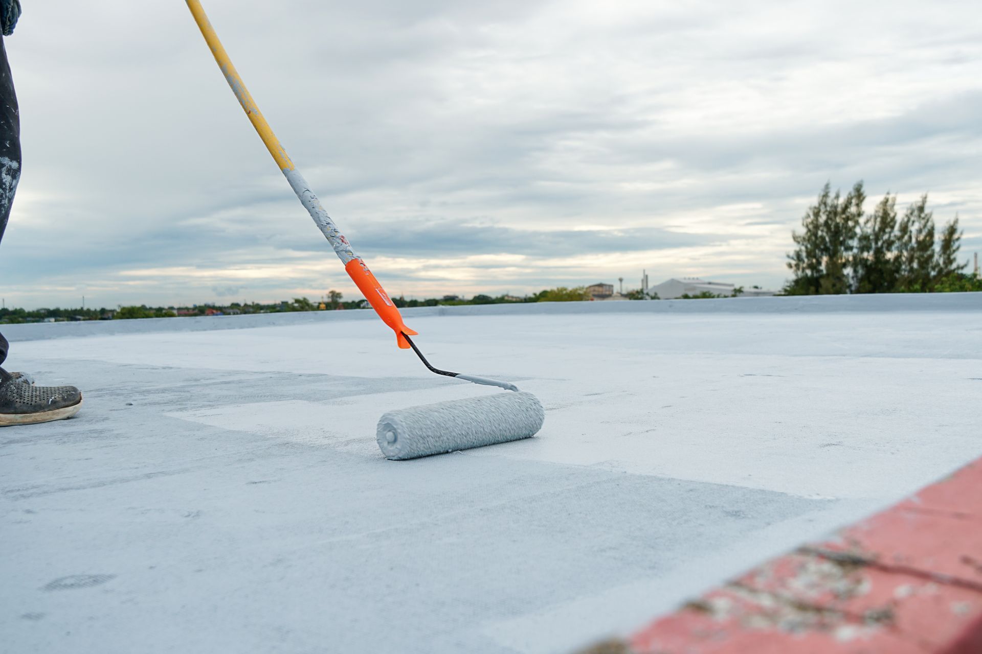 Person applying a white protective coating to a flat roof surface with a paint roller.