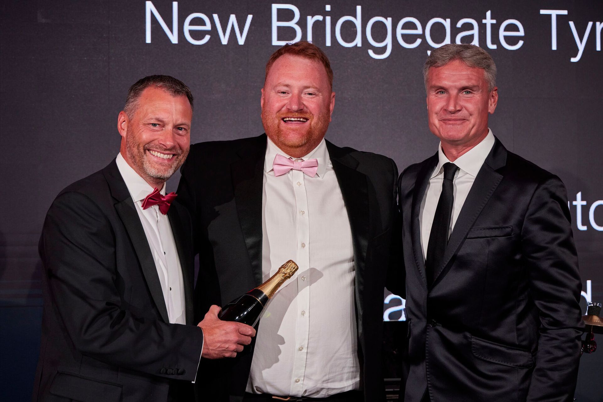 Three men in suits are posing for a picture and one of them is holding a bottle of champagne.