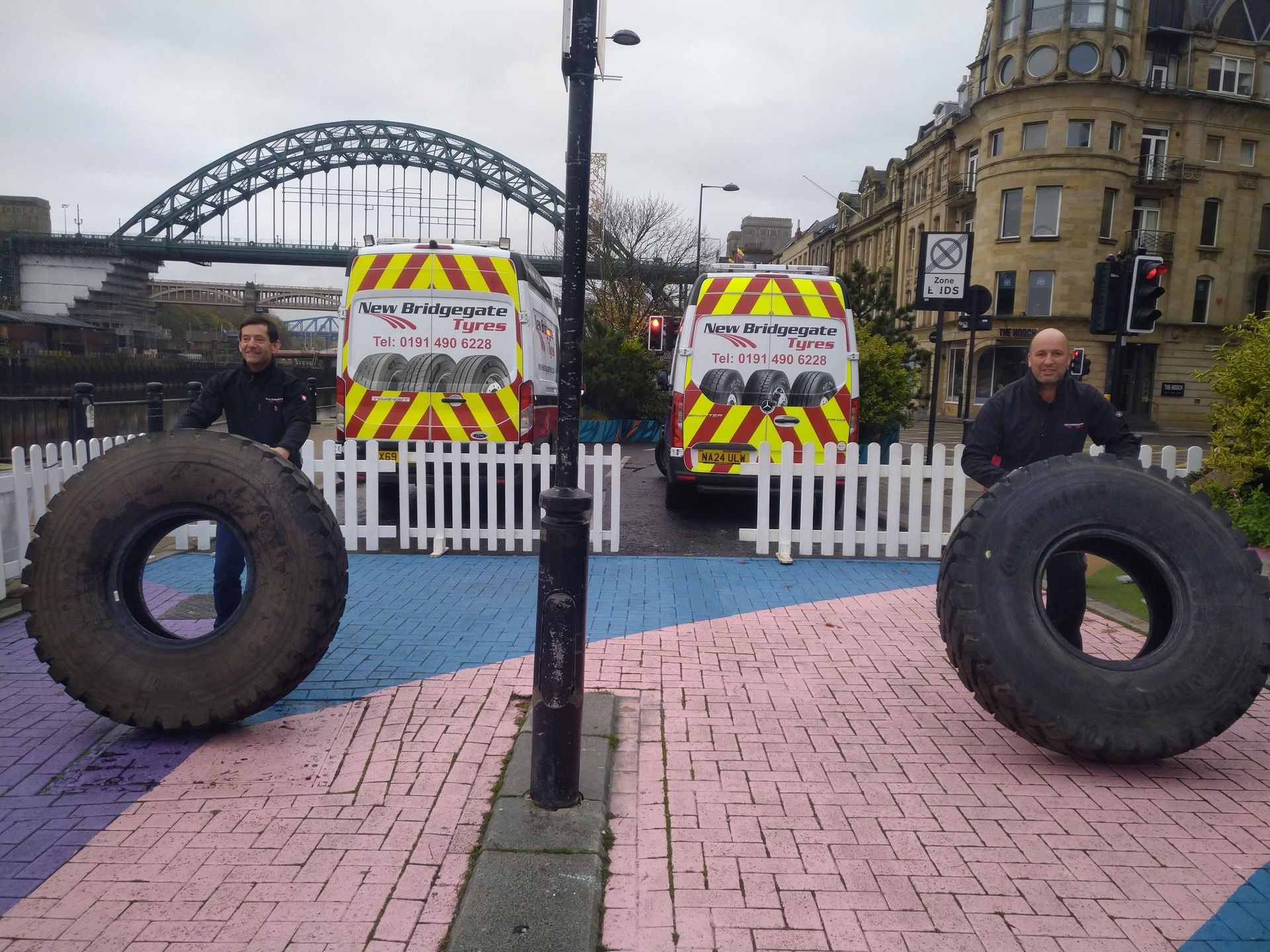Two men holding large tires in front of a van that says ' tires ' on it