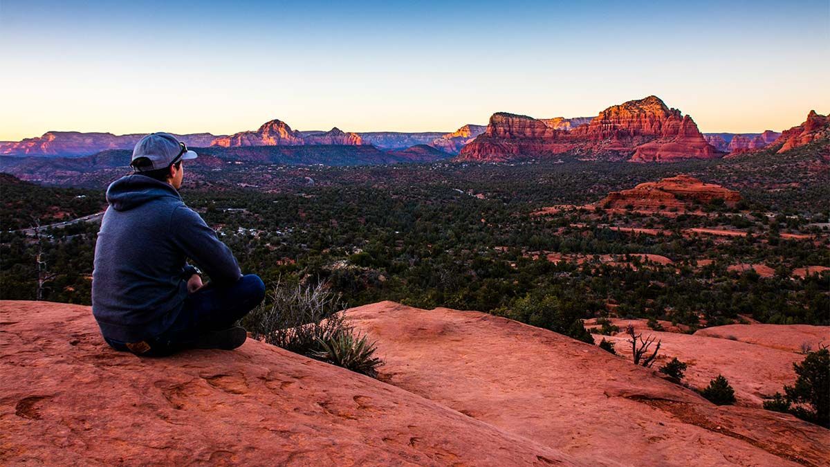 Person seated on a red rock overlooking a lush valley and distant red rock formations at sunset.
