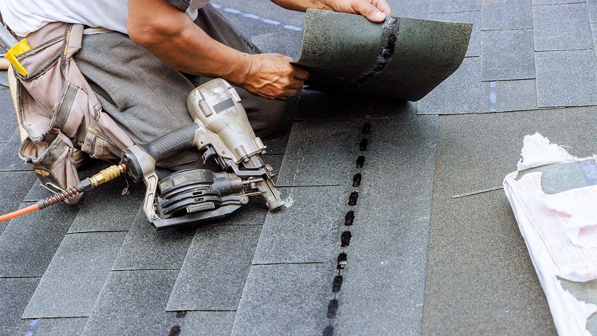 Roofer kneeling on a roof, installing a shingle with a nail gun.